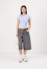Top cropped justo azul claro, calções de ganga largos cinzentos, ténis brancos e uma pequena bolsa branca. Estilizado de forma casual contra um fundo simples.