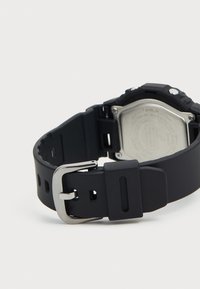Bracelet de montre en caoutchouc noir avec une boucle en métal argenté. L'arrière de la montre présente un boîtier circulaire en acier inoxydable et des détails gravés.