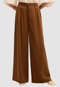 Pantaloni larghi marroni con vita alta, caratterizzati da pieghe, chiusura con bottone e una texture liscia e morbida. Completati da scarpe chiare.