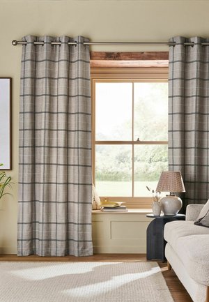 Beige living room with window framed by plaid curtains, beige sofa, black side table with lamp, and books on window seat.