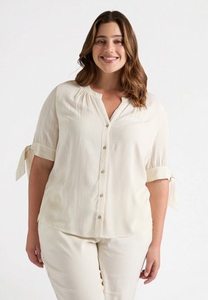 Blusa di colore crema con scollo a V, chiusura a bottoni, tessuto plissettato e polsini arrotolati legati con fiocchi. Tessuto liscio e vestibilità rilassata.