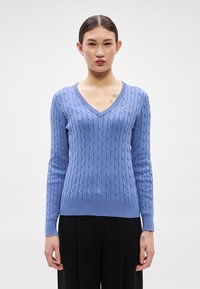 Gaiši zils džemperis ar V veida apkakli, pītas adīšanas rakstu, ribotām aprocēm un apakšu, ar mazu izšūtu detaļu kreisajā krūšu daļā.