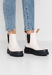 Botas Chelsea blancas con suelas negras y paneles laterales elásticos. Presentan una lengüeta anudada de color naranja. Suela de goma texturizada para agarre.