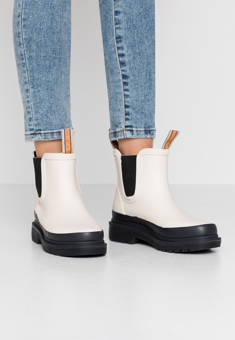 Botas Chelsea blancas con suelas negras y paneles laterales elásticos. Presentan una lengüeta anudada de color naranja. Suela de goma texturizada para agarre.