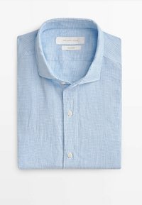Chemise en lin bleu clair pliée avec motif à carreaux blancs et col boutonné sur fond blanc.