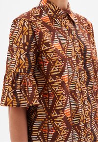 Camicia corta marrone a maniche corte con una varietà di motivi geometrici in arancione, giallo e nero, dotata di bottoni frontali e maniche arricciate.