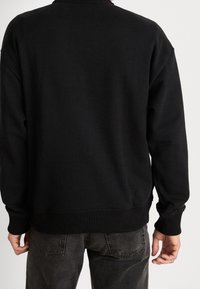 Uomo che indossa un maglione nero con orlo a costine e jeans scuri, visto da dietro su uno sfondo chiaro e uniforme.