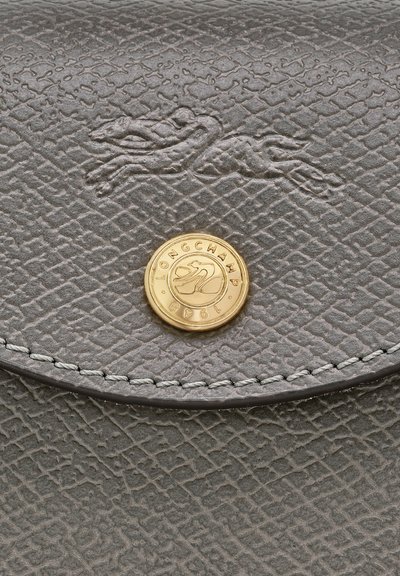 Graues strukturiertes Lederklappfach mit eingeprägtem laufendem Pferdelogo und rundem goldenen Knopf, der mit "Longchamp 1948" und Pferdemotiv graviert ist.
