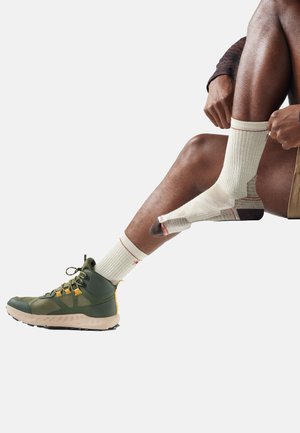 Person adjusting beige and brown socks while wearing green hiking shoes and brown shorts, seated against a white background.