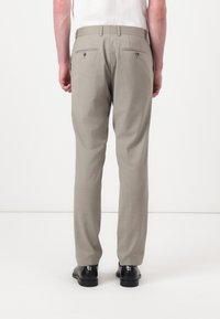 Calças bege com um corte ajustado, apresentando duas bolsas traseiras com fechos de botão e uma textura suave, combinadas com sapatos pretos.
