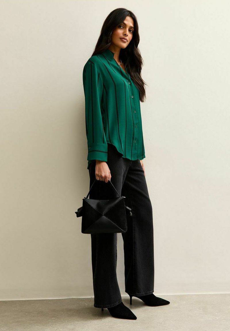 Groen blouse met verticale zwarte strepen, gecombineerd met zwarte wijde broek en een zwarte geometrische handtas. Zwarte puntige schoenen.