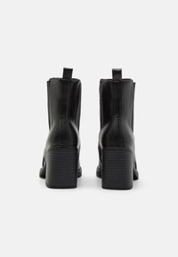 Botas de tobillo de cuero negro con una textura suave, tacón elevado, detalle de cremallera y tirador. Cuenta con una punta redondeada y suela resistente.