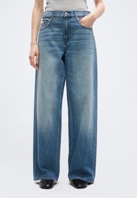 Άτομο που φοράει μπλε τζιν παντελόνι ψηλόμεσο με φαρδιά μπατζάκια και ξεθωριασμένο σχέδιο, μαζί με μαύρα παπούτσια με τετράγωνη μύτη, στέκεται με το ένα χέρι στην τσέπη.