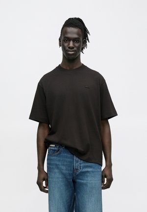 Hombre joven con cabello oscuro, vestido con una camiseta negra de manga corta y jeans azules, de pie contra un fondo blanco liso.