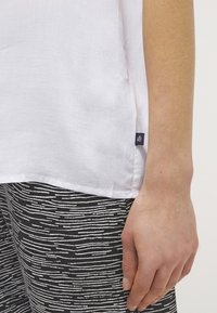 Detailní záběr na ruku vedle bílé lněné blůzy s malou černou visačkou a černými kalhotami s nepravidelnými bílými horizontálními pruhy.