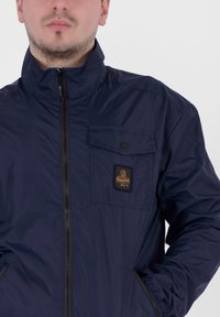 La helina azul con cremallera está hecha de tejido ligero. Presenta un bolsillo en el pecho con cierre de botón y un parche de logo. Textura suave y cuello alto.