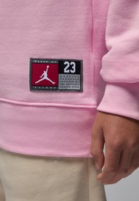 Rosa genser med ribbestrikkede mansjetter, med en svart og rød lapp som viser "Jordan"-logoen, nummer 23, og teksten "Brand of Flight".