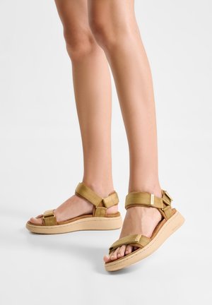 Benen met beige platformsandalen met verstelbare bandjes over tenen, enkels en hakken op een effen witte achtergrond.