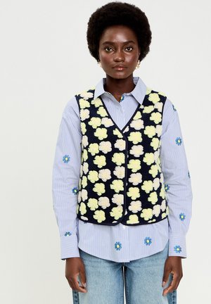 Donna che indossa un gilet blu navy con motivi floreali color crema sopra una camicia a righe blu con fiori ricamati blu, abbinata a jeans azzurri.