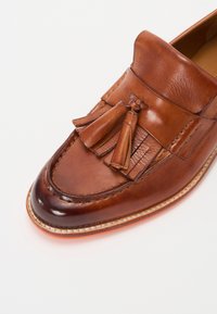 Mocasin de cuero marrón con acentos de borlas, puntera pulida y costuras detalladas a lo largo de la suela. Textura suave con un diseño clásico.