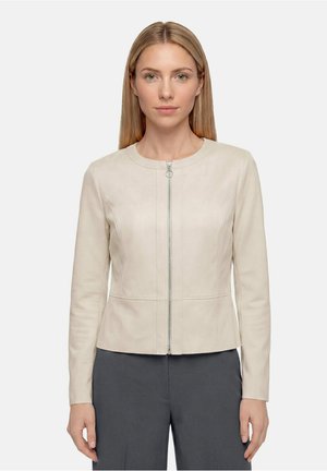 Femme aux cheveux blonds et raides, portant une veste beige zippée et un pantalon gris foncé, regardant devant elle sur un fond uni.