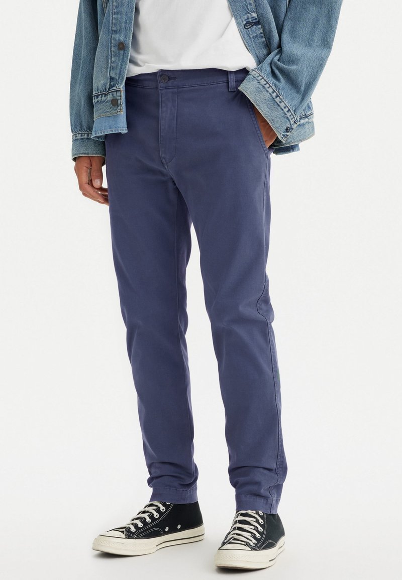 Chinos de algodón azul marino con un corte ajustado, bolsillos delanteros y cierre de botón, combinados con zapatillas negras de caña alta y una chaqueta de mezclilla.