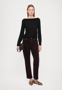 Schwarzes, schulterfreies Oberteil, burgunderfarbene Cord-Hose und Loafers im Leopardenmuster. Das Model hält eine kleine schwarze Handtasche. Weißer Hintergrund.