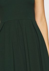 Robe vert foncé avec un corsage ajusté et une jupe plissée. Fabriquée dans un tissu doux et texturé. Le design présente une couture subtile à la taille.