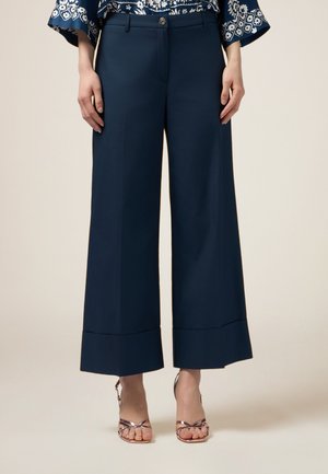 Pantalons larges en navy avec une texture lisse, dotés d'une fermeture à bouton et d'un ourlet évasé. Associés à des sandales métalliques à lanières.