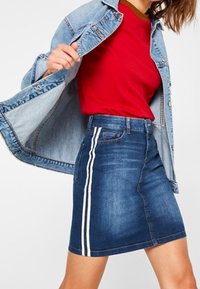 Minifalda de mezclilla en azul, con un ajuste ceñido y rayas laterales blancas; combinada con una camiseta de cuello redondo roja y una chaqueta de mezclilla clara.