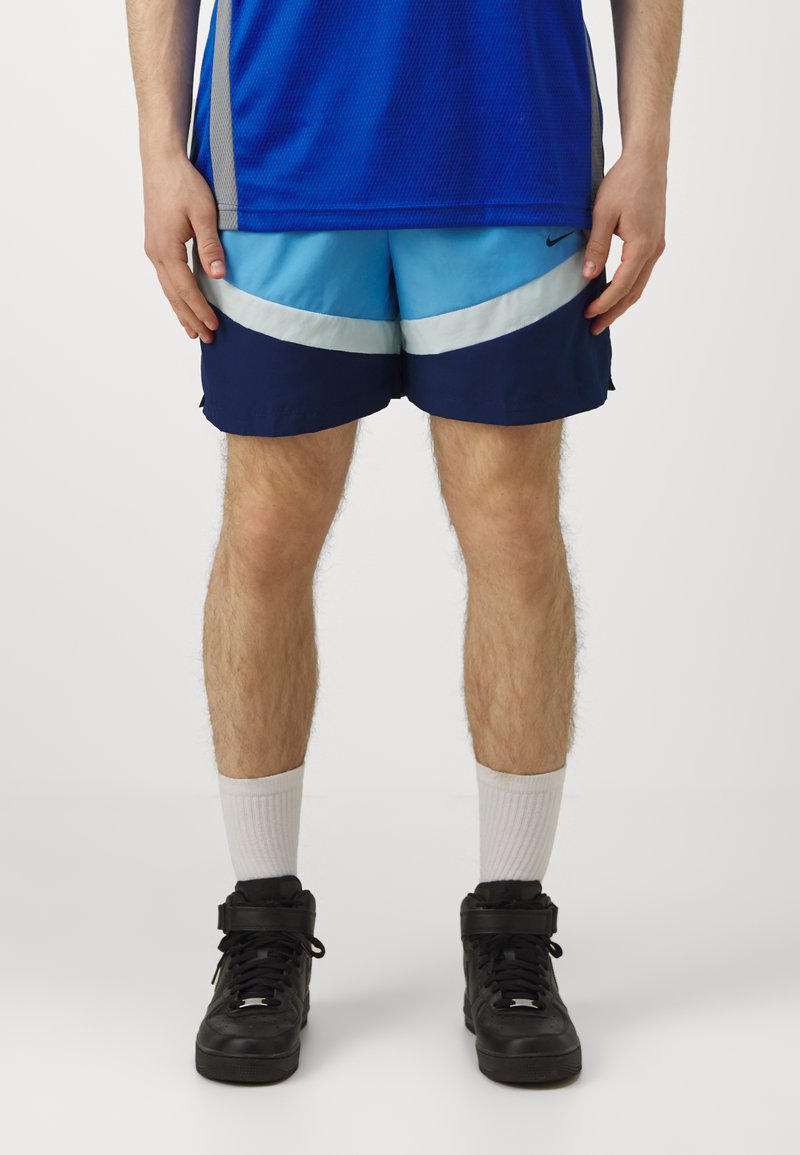 Pantaloni corti blu e navy con strisce bianche, realizzati in materiale leggero. Abbinati a calzini bianchi alla caviglia e sneakers nere alta.