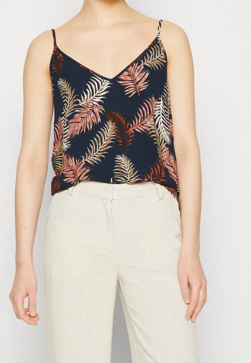 Laivastonsininen olkaimeton toppi, jossa on lehväkuvio vaaleanpunaisen, kermanvärin ja kullan sävyissä, valmistettu kevyestä kankaasta, yhdistettynä beigeihin housuihin.