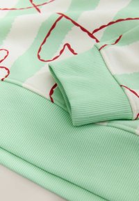 Világoszöld pulóver piros firkamintákkal; bordázott mandzsettákkal és szegéllyel. Az anyag sima textúrát mutat kontrasztos csíkokkal.