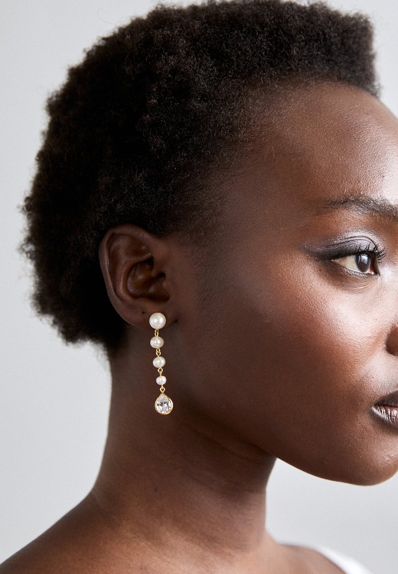 Χρυσές κρεμαστές σκουλαρίκια που διαθέτουν τρεις λευκές πέρλες και ένα διαφανές κρυστάλλινο δάκρυ στο κάτω μέρος, παρουσιασμένα σε ουδέτερο φόντο.