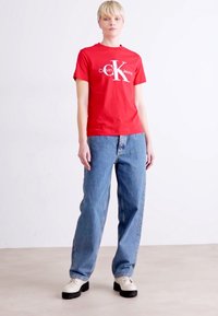 Rode katoenen T-shirt met wit "Calvin Klein Jeans" logo, gecombineerd met lichtblauwe, losse denim jeans en witte schoenen, staande op een neutrale achtergrond.
