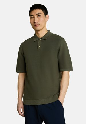 Man wearing an olive green short-sleeve knit polo shirt with two buttons, standing against a plain light background.