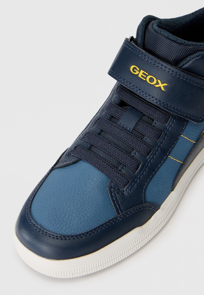 Sneaker sintetico blu navy con finitura in pelle testurizzata, lacci elastici e cinturino in velcro. Logo dorato "GEOX" sul cinturino. Suola in gomma bianca.