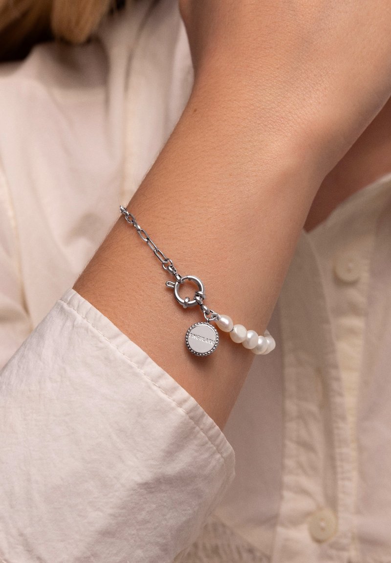 Pulsera de cadena de plata con acentos de perlas blancas y un charm redondo con texto grabado, llevada en una muñeca de tono de piel claro.