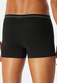 Bakifrån av en person som bär svarta sömlösa boxerbriefs med en subtil grå rand på midjebandet mot en vit bakgrund.