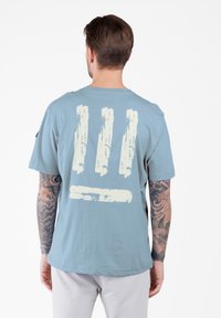 Mann mit tätowierten Armen, der ein hellblaues T-Shirt mit vier weißen Pinselstrich-Designs auf dem Rücken trägt und vor einem weißen Hintergrund steht.