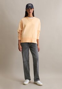 Sweat-shirt pêche clair à col rond, poignets côtelés, associé à un jean gris droit avec des accents usés et des baskets blanches.