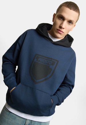 Młody mężczyzna z krótkimi włosami, ubrany w granatową bluzę z kapturem z czarną podszywką i delikatnym logo tarczy "PROSTO", ręce w przedniej kieszeni.