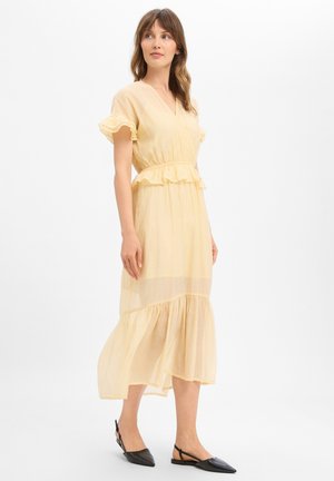 Woman wearing a pale yellow midi dress with ruffled sleeves and hem, standing and looking to the side, wearing black pointed flat shoes.