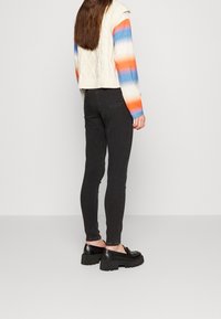 Jeans skinny negros combinados con un suéter corto de punto trenzado en color crema, que presenta rayas horizontales en azul, naranja y blanco, y zapatos de tacón grueso negros.