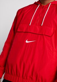 Rode nylon pulloverjack met een voorzak, ritskraag en witte trekkoorden. Bevat een wit Nike-logo op de borst. Gelaagde afwerking.