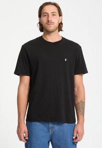 Camiseta de algodón negra con cuello redondo, mangas cortas y un pequeño logo blanco en el lado izquierdo del pecho. Combinada con unos pantalones vaqueros azules.