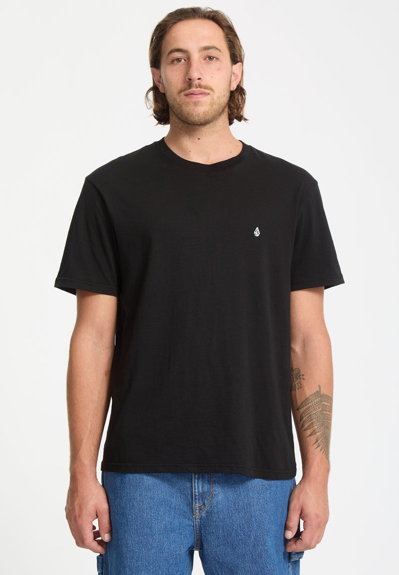 Camiseta de algodón negra con cuello redondo, mangas cortas y un pequeño logo blanco en el lado izquierdo del pecho. Combinada con unos pantalones vaqueros azules.