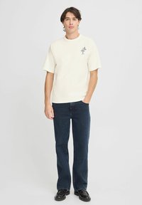 Cremefarvet t-shirt med korte ærmer og et blomstermotiv på venstre bryst, parret med mørkeblå lige bukser og sorte sko.