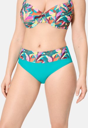Mujer con un traje de baño de dos piezas con parte inferior turquesa y parte superior y cinturilla con estampado colorido de hojas tropicales.