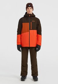 Skijack in bruin en oranje met blokachtige kleurontwerp, ritszakken, elastische manchetten, gedragen over olijfgroene broeken en zwarte laarzen.